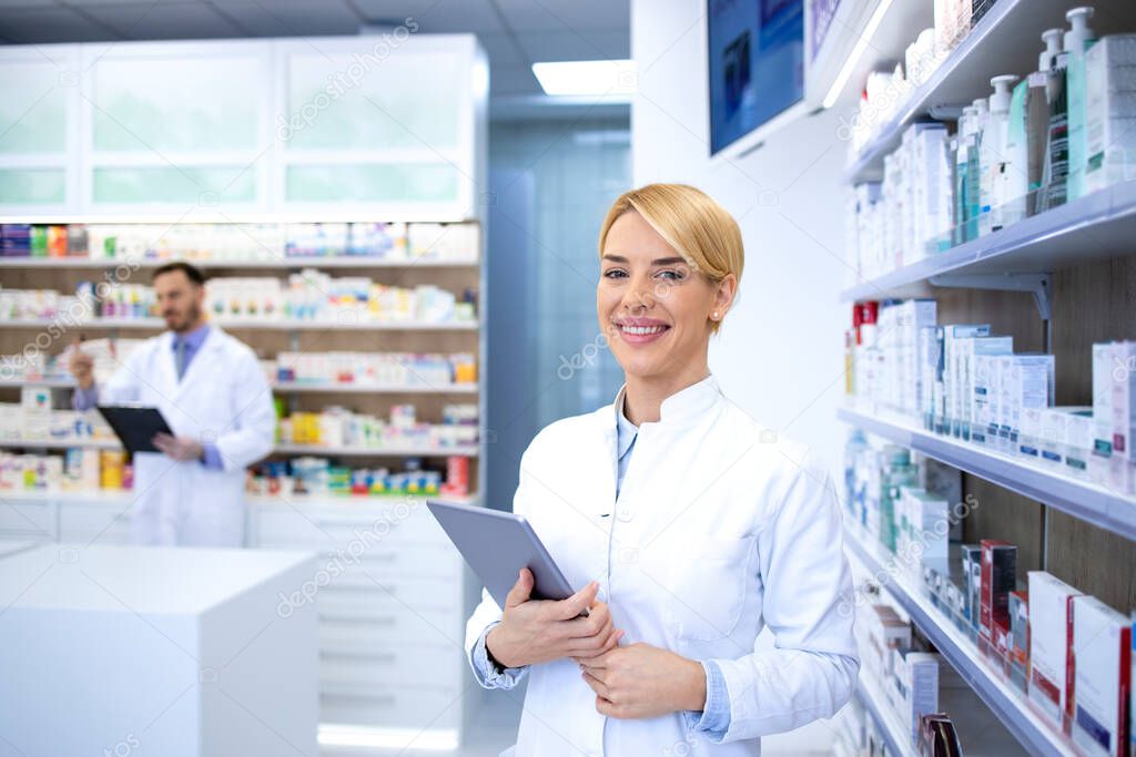 Portrait of smiling female blonde pharmacist standing in pharmacy shop or drugstore by the shelf with medicines and holding tablet.