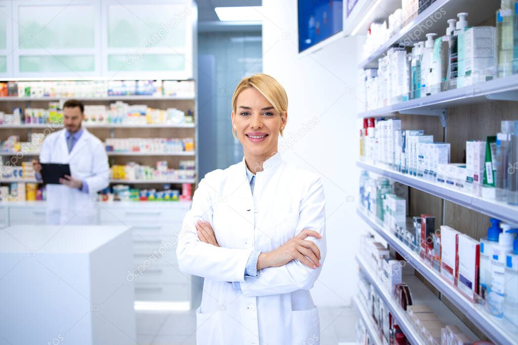 Portrait of beautiful female blonde pharmacy worker standing in drugstore with her arms crossed. In background shelves with medicines.