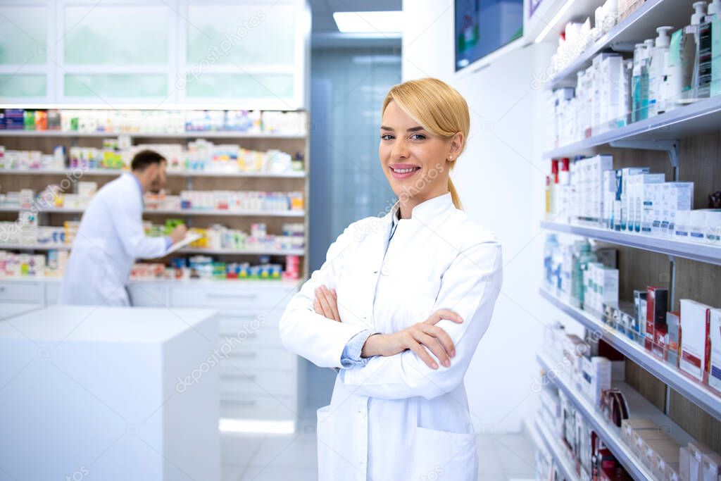Portrait of smart looking female blonde pharmacist standing in pharmacy shop or drugstore with her arms crossed. In background shelves with medicines.