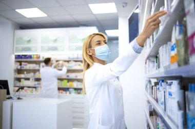 Kovid-19 virüs salgını sırasında eczanede elinde ilaç tutan yüz maskesi ve beyaz önlük giyen kadın eczacı..