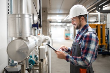 Sanayi mühendisi elinde tablet bilgisayar tutuyor ve fabrika kazan dairesinde ısıtma sisteminin parametrelerini ayarlıyor..