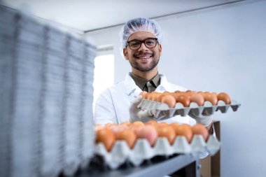Yiyecek fabrikası işçisi, beyaz önlüklü, saç filesi takmış ve temizlik eldivenleriyle ambalaj makinesinin yanındaki yiyecek işleme tesisinde yumurta kasası tutuyor..