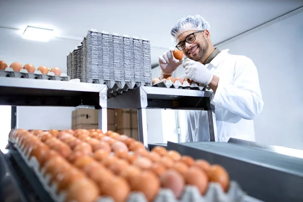 Technologist in white coat and hairnet checking quality of industrially produced eggs in food processing plant or farm.