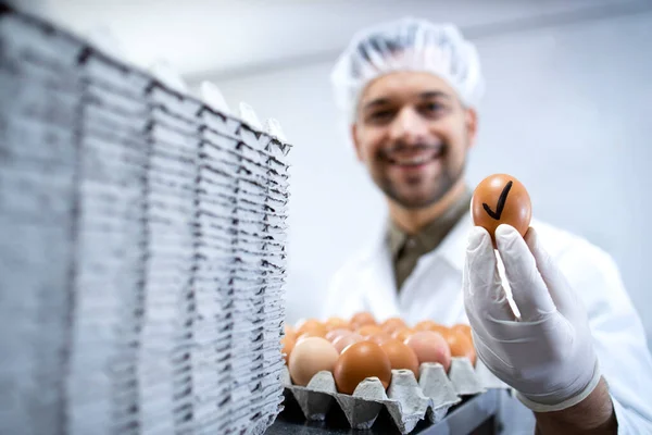 Food factory technologist standing by industrial eggs sorting machine and holding an egg that passed quality control test.