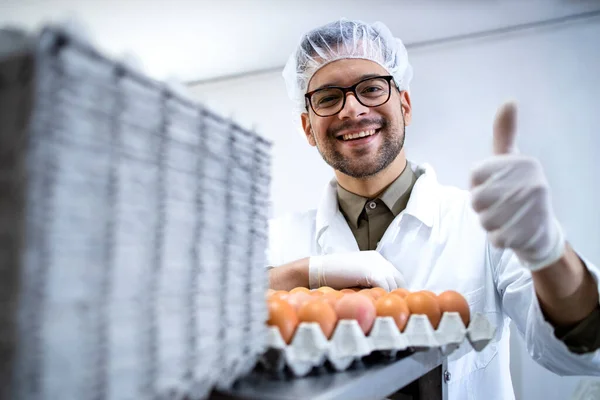 Food factory technologist standing by industrial eggs packaging machine and holding thumbs up.