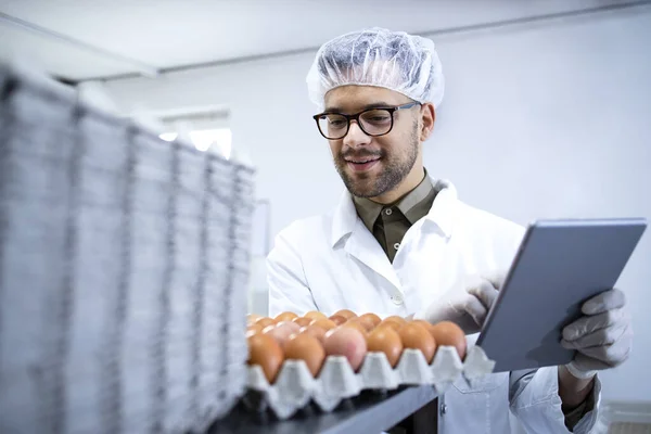 Food factory worker in white coat hairnet and hygienic gloves controlling eggs production at the food processing plant on tablet computer.