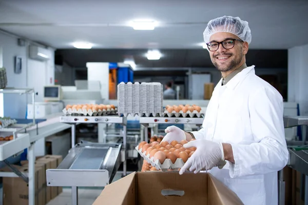 Food factory worker in white coat hairnet and hygienic gloves packing eggs in cardboard box preparing them for the market.