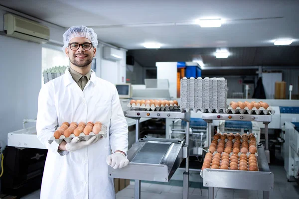 Portrait of food factory worker with hairnet and hygienic gloves holding eggs by industrial transporting and packing machine in food processing plant.