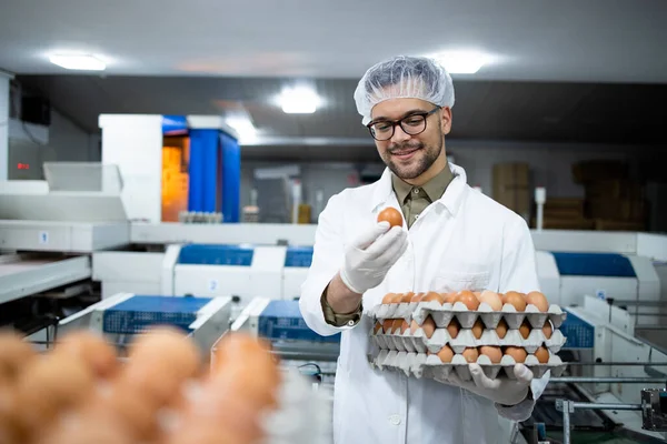 Portrait of food factory worker with hairnet and hygienic gloves checking quality of eggs by industrial transporting and packing machine in food processing plant.