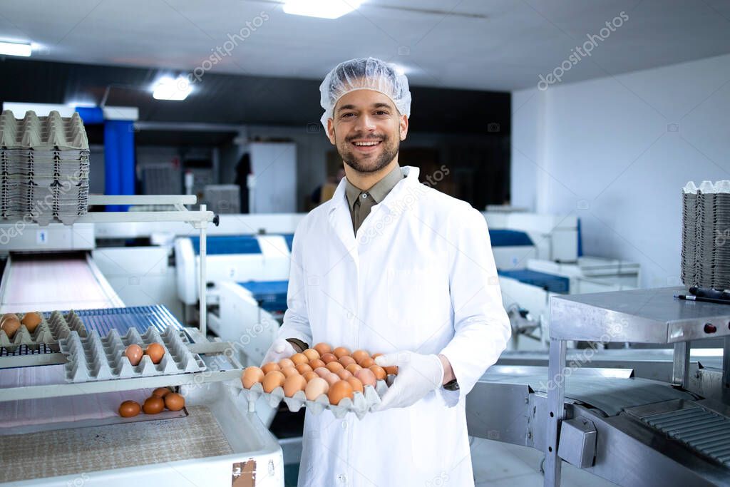 Portrait of an egg farm worker holding cardboard crate with fresh eggs. In background machine transporting eggs for packaging.