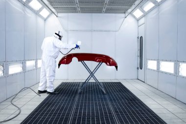 Car painter in protective clothes and mask painting automobile bumper with metallic paint and varnish in chamber workshop. clipart