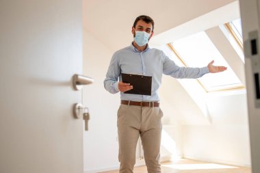 Portrait of male real estate agent with face mask standing in new house during corona virus pandemic.
