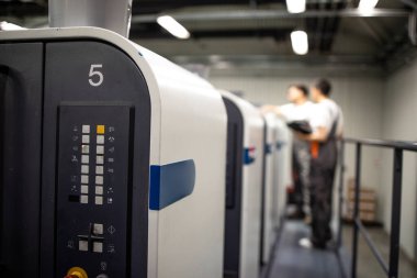 Yazdırma işleminin kalitesini ve kontrol sürecini kontrol eden çalışma üniformasında çalışan modern ofset yazdırma makinesi ve operatörlerinin görünümünü kapat.