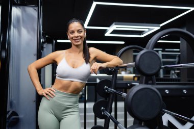 Spor salonunda ağırlık kaldırma aletlerine yaslanan profesyonel kadın antrenör portresi..