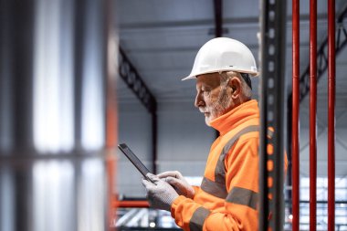 Başlık ve güvenlik yeleği giyen kıdemli fabrika işçisi dijital tablet verilerini kontrol ediyor.