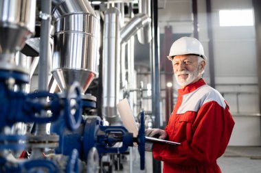 Fabrikada dizüstü bilgisayar kullanarak üniforma ve şapka takan olgun bir mühendis.