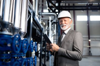 Sert şapkalı kıdemli işadamı endüstriyel ekipmanları kontrol ediyor, gülümsüyor