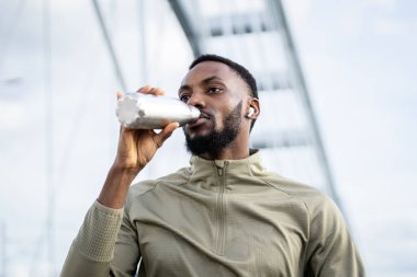 Genç siyahi bir adam açık hava antrenmanı sırasında susuz kalıyor. Yeniden kullanılabilir bir su şişesinden içmenin tadını çıkarıyor.