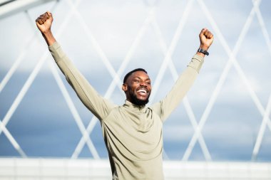 Spor giyim sektöründe genç ve mutlu bir zenci. Yumruklarını sıkmış ve gülümsüyor. Açık havada idman yaptıktan sonra başarıyı kutluyor.