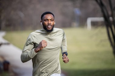 Genç atletik siyah adam spor yapıyor, parkta koşuyor spor kıyafetleri giyiyor ve kulaklık takıyor.