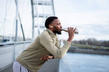 Afrika kökenli Amerikalı erkek koşucu, yoğun bir açık hava eğitiminden sonra su şişesiyle su içmek için köprüde duruyor.
