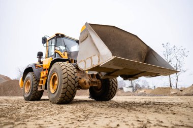 Sarı tekerlek yükleyicisi büyük ön kovasını yerleştiriyor, ağır toprak taşıma görevlerine hazır, inşaat alanında ya da taş ocağında malzeme toplamaya hazırlanıyor.