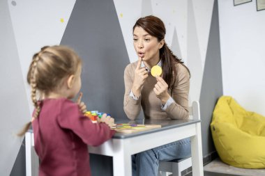 Çocuklu bir konuşma terapisti bir masada öğrenim etkinliği içinde, destekleyici bir eğitim ortamında sözlü beceri pratiği ve konuşma geliştirmeye odaklanıyor.