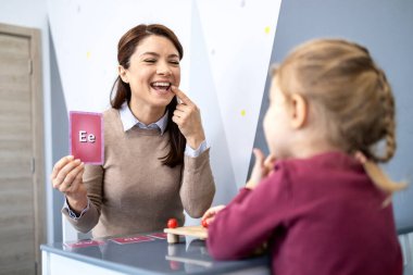 Konuşma terapisti genç bir kızla klinikte çalışıyor, telaffuz için düzgün bir ağız pozisyonu sergiliyor ve dil dersi sırasında elinde bir alfabe bilgi kartı tutuyor.