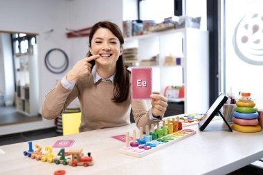 Konuşma terapisti, neşeli bir klinik ortamında oyuncaklarla konuşma, kelime hazinesi ve iletişim becerileri geliştirmek için bir çocuğa fonik ve alfabe bilgi kartları aracılığıyla rehberlik ediyor.