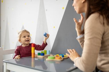 Genç bir çocuk renkli ahşap bloklarla öğrenme aktivitesine girişiyor, bir yetişkine bakıyor, iyi motor beceriler ve konuşma terapistiyle iletişim kuruyor.