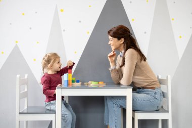 Anaokulu öğretmeninin gülümseyen bir çocukla etkileşimi, küçük bir masada renkli ahşap yapı taşlarıyla oynaması, yaratıcı bir anaokulu ortamında erken öğrenim ve gelişimi teşvik etmesi.