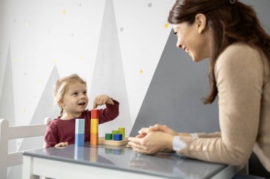 Bir masada renkli ahşap bir kule inşa eden bir kıza rehberlik eden anaokulu öğretmeni motor becerilerini, yaratıcılığını, konsantrasyonunu ve çocuk odasında eğlenceli erken öğrenimi teşvik ediyor.