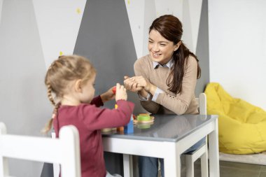 Anaokulu masasında eğitici tahta bloklarla oynayan ve onunla etkileşim içinde olan gülümseyen anaokulu öğretmeni bağlanma ve gelişimsel öğrenmeyi destekliyor.