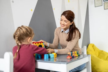 Çocukların zihinsel ve motor beceri gelişimini desteklemek için şekil ayırıcı ve yapı taşları kullanan, oyun zamanı boyunca küçük bir kızla takılan neşeli anaokulu öğretmeni.