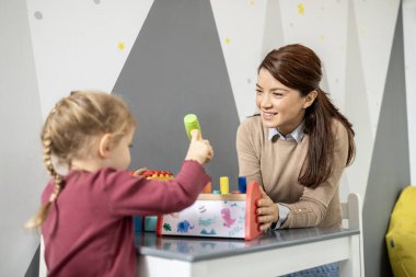 Anaokulu öğretmeni gülümseyen ve renkli tahta çekiç oyuncağıyla oynayan küçük bir çocuğu seyreden, erken eğitimi, motor becerilerini geliştirmeyi ve anaokulunda yaratıcı oyunları teşvik eden,