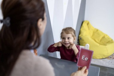 Konuşma terapisti, gülümseyen bir anaokulu kızına alfabe bellek kartıyla konuşma geliştirme, güven ve erken dil becerilerini klinikte teşvik ederek eklem egzersizleri yaptırıyor.