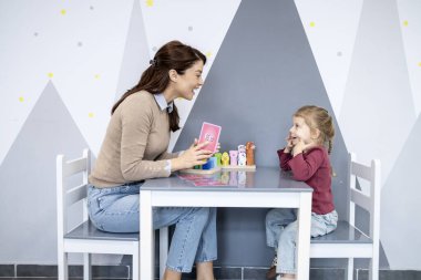 Dişi konuşma terapisti, parlak dekore edilmiş bir odada küçük bir kızla takılıyor, konuşma, fonik ve iletişim gelişimine odaklanmış bir eğitim aktivitesinde ona rehberlik ediyor.