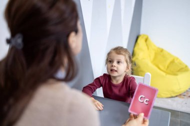 Konuşma terapisti, okuma yazma ve öğrenme becerileri üzerine odaklanan bir eğitim oturumu sırasında dil gelişimi ve iletişim konusunda bir hafıza kartı kullanan küçük bir çocuğa yardım ediyor.