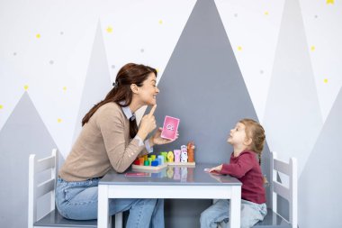 Genç bir kızla dil geliştirme üzerine çalışan kadın konuşma terapisti, eğitim oyunları ve alfabe araçları kullanarak konuşma düzeltme ve iletişim becerilerine odaklanıyor