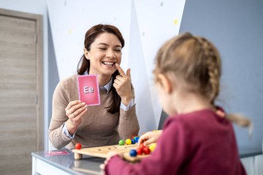 Konuşma terapisti bir dil geliştirme oturumunda küçük bir çocukla ilgileniyor, elinde e harfiyle bir hafıza kartı tutuyor ve ses üretimi için düzgün bir ağız eklemlerini gösteriyor.
