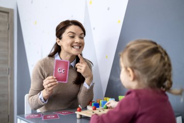 Konuşma terapisti, küçük bir çocuğa ağız hareketleri ve alfabe bilgi kartlarıyla telafuz, eklem ve iletişim becerilerini geliştirmek için neşeli bir sınıfta rehberlik ediyor.