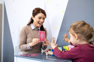 Konuşma terapisti, dil, sayma ve iletişim becerilerini geliştirmek için eğlenceli bir oturum sırasında gülümseyen bir anaokulu öğrencisini numara kartlarıyla ve oyuncaklarla meşgul ediyor.