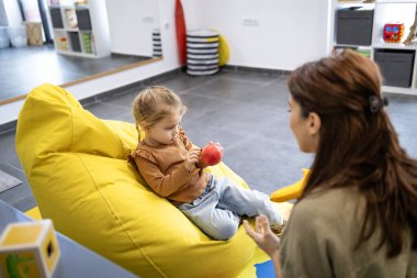 Renkli bir odada terapi seansı sırasında küçük bir kızla ilişki kuran çocuk psikoloğu, bir fasulye torbasının üzerine oturmuş, bir elma tutuyor, zihinsel ve duygusal gelişimine odaklanıyor.