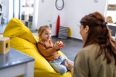 Bir çocuk psikoloğuyla terapi seansı sırasında gülümseyen ve etkileşen küçük bir kız, rahat ve destekleyici bir ortamda duygular ve duygusal gelişimi tartışıyor.