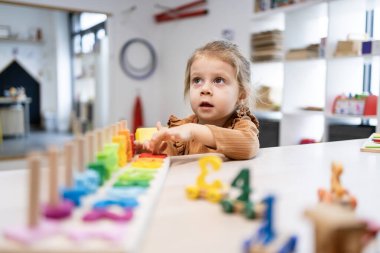 Anaokulu öğrencisi parlak bir sınıf masasında renkli ahşap oyuncaklar ve bulmacalar üzerinde yoğunlaşıyor, iyi motor beceriler geliştiriyor, sayıyor, erken matematik kavramları ve eğlenceli öğrenimler geliştiriyor.