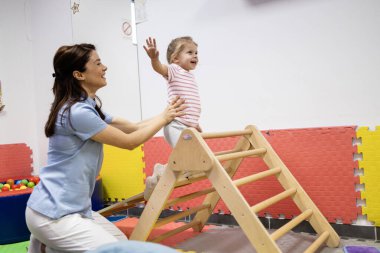 Uzman bir pediatrik terapist, yürümeyi öğrenen küçük bir kıza yardım ediyor ve mesleki terapi seansı sırasında içeride bir üçgen üzerinde vücudunu güçlendiriyor.