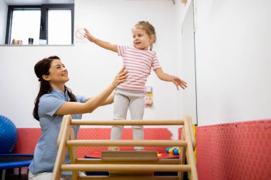 Çocuk doktoru, yüzü gülen bir çocuğa yardım ediyor tahta bir yapıya basıyor, motor gelişimi ve denge eğitimi için fizik tedavi egzersizleri yapıyor.