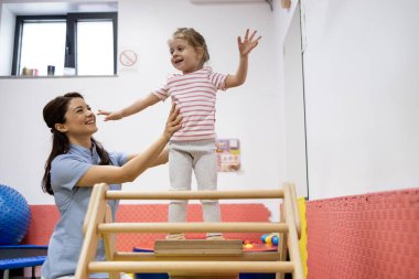 Gülümseyen pediatrik meslek terapisti egzersiz aparatı üzerinde dengede duran bir çocuğu destekliyor, bir rehabilitasyon kliniğinde motor gelişimini teşvik ediyor ve kas gücünü artırıyor.