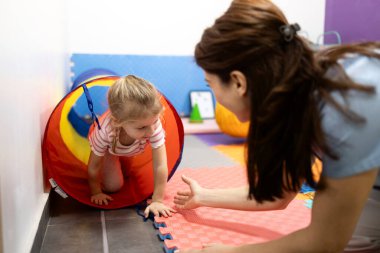 Genç kız yumuşak paspasların üzerinde renkli bir oyun tünelinde sürünüyor. Rehabilitasyon sırasında kadın bir pediyatrik terapistin rehberlik ettiği motor beceriler ve fiziksel gelişime odaklanmak