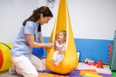 Meslek terapisi sırasında renkli duyusal entegrasyon salıncağında gülümseyen bir kıza rehberlik eden kadın pediatrik terapist motor becerileri, dengeyi, koordinasyonu ve neşeli rehabilitasyonu teşvik ediyor.